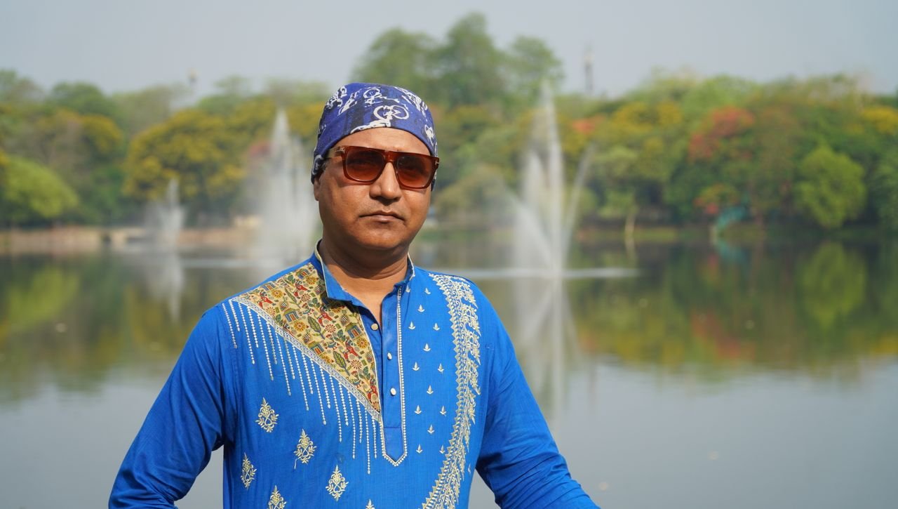 A man posing by a lake in Jamshedpur, India, wearing a blue kurta and sunglasses.